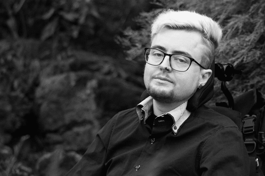 A black and white image of Jamie Hale. They are white, and surrounded by nature, wearing a black shirt, with black glasses frames and bleached blonde hair.