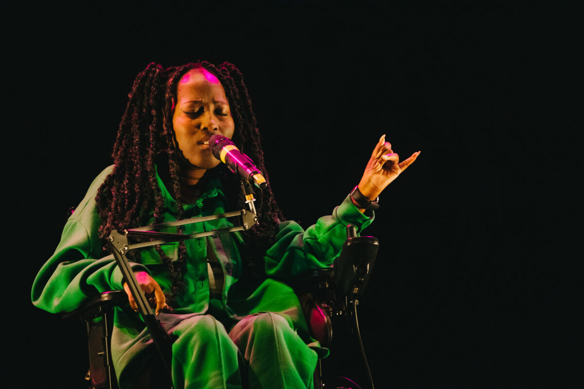Jacqui is a black woman with black waist-length twists, She is wearing a green velvet tracksuit. She is sitting in her powered wheelchair and singing into a microphone with her eyes closed.