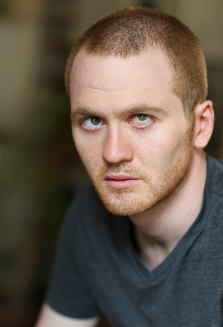 A headshot of Sam Brewer, a white man with blue eyes, short blonde hair and beard. Sam looks softly into the camera. He is wearing a blue t-shirt.