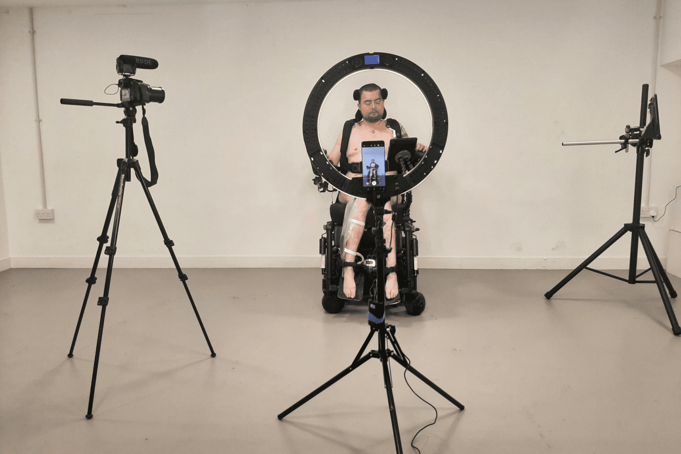 Jamie Hale sits naked in their electric wheelchair, surrounded by cameras on tripods. Jamie is a white person with red hair cropped in a buzz cut. Their black wheelchair harness straps and catheter bag are visible behind the cameras.