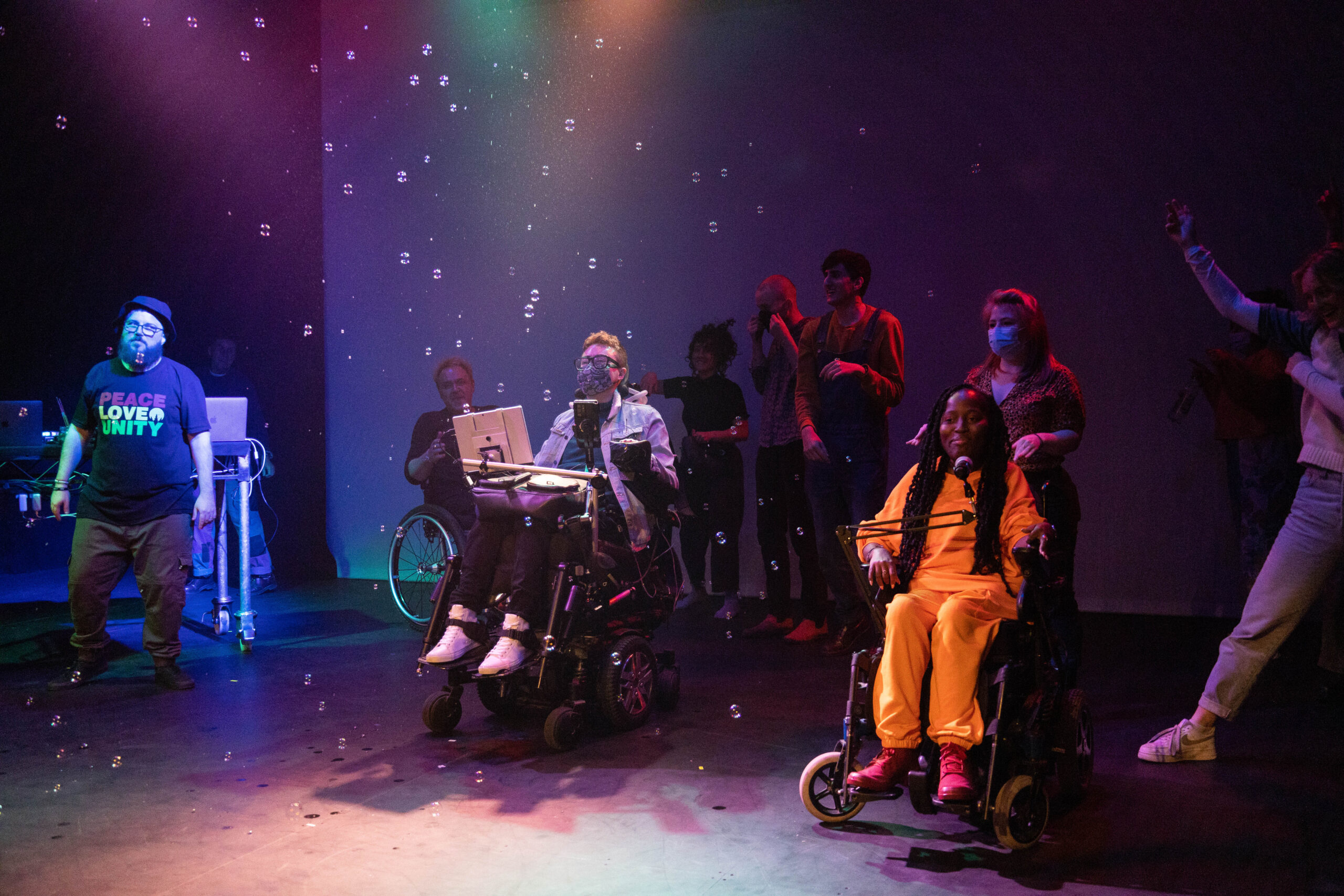The cast & crew of the 2021 CRIPtic Showcase gather on stage for a final bow. They are bathed in blue and pink light and surrounded by bubbles.