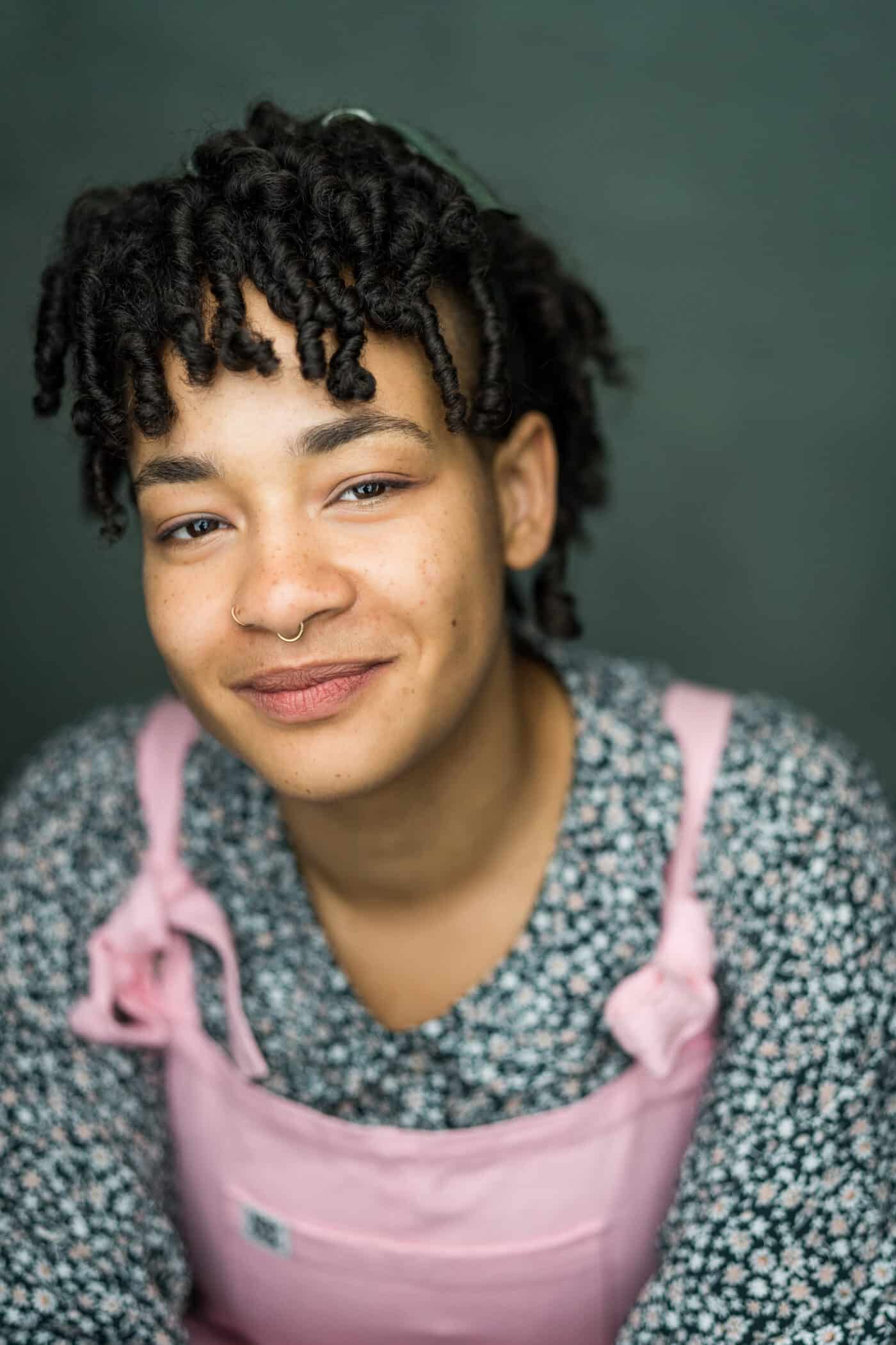 A lighter skinned Black non-binary person in pink dunagrees and a dark green floral shirt leans towards the camera. They are smiling and have ringlets covering their forehead.