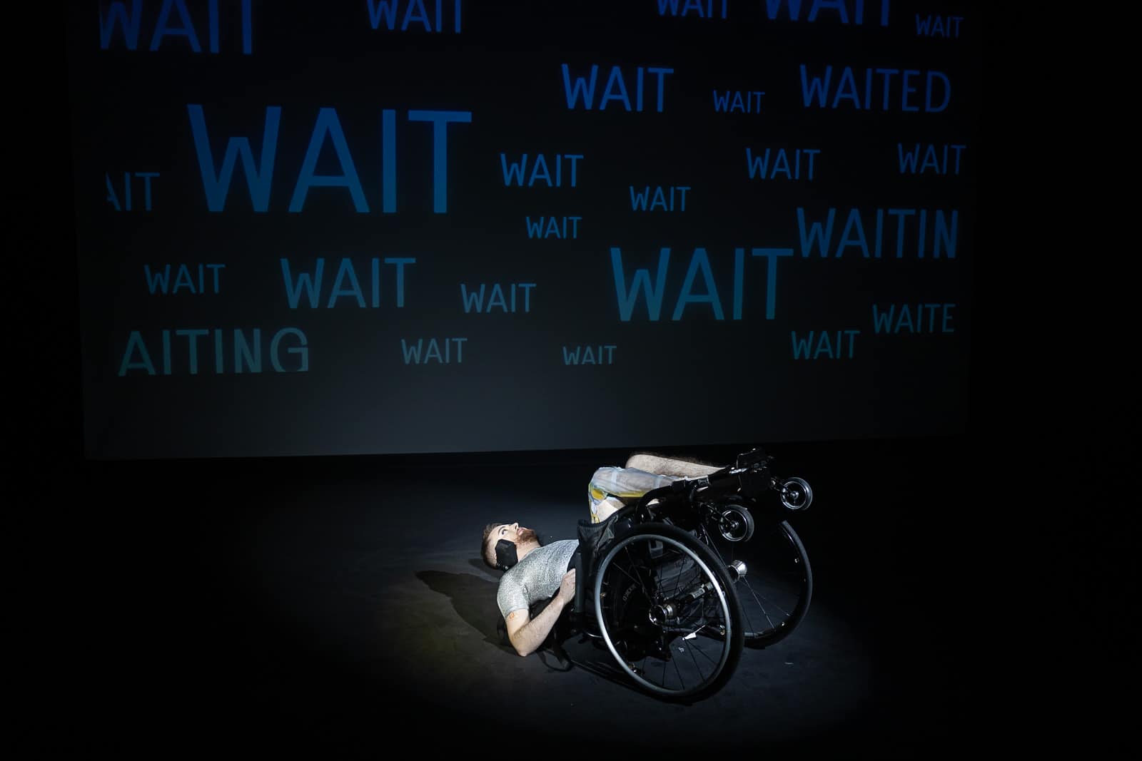 Jamie Hale performs NOT DYING. Jamie, white person with red hair is laid on the floor in their manual wheelchair. The are wearing a silver shirt, maroon boxers and their catheter bag is seen on their right leg. Behind them, the word 'WAIT' is projected repeatedly across the back wall of the theatre.
