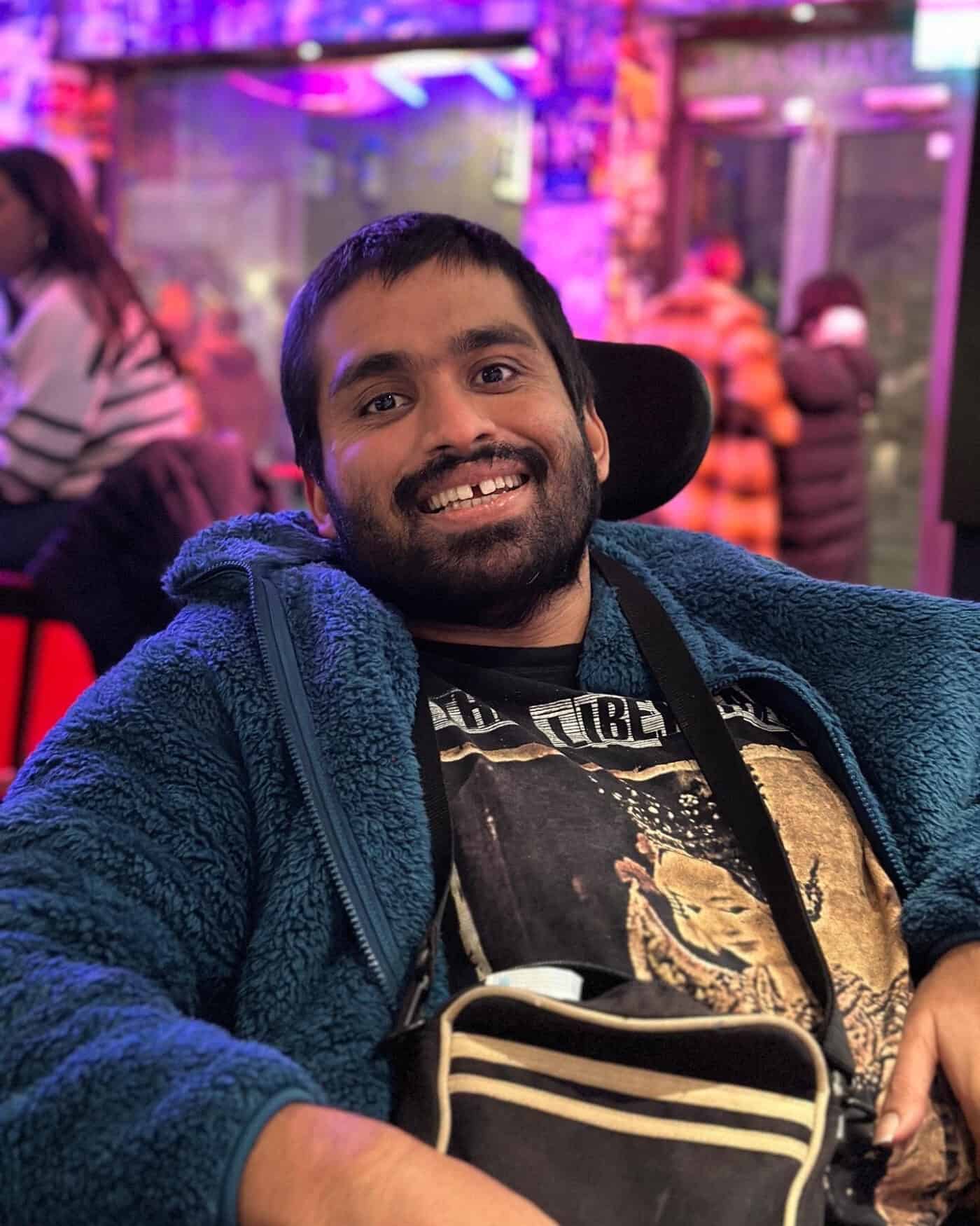 Photo of Jamil Dhillon. Jamil is wearing a blue fluffy jacket and black t shirt sitting in his powerchair with a small manbag slung over his left shoulder.