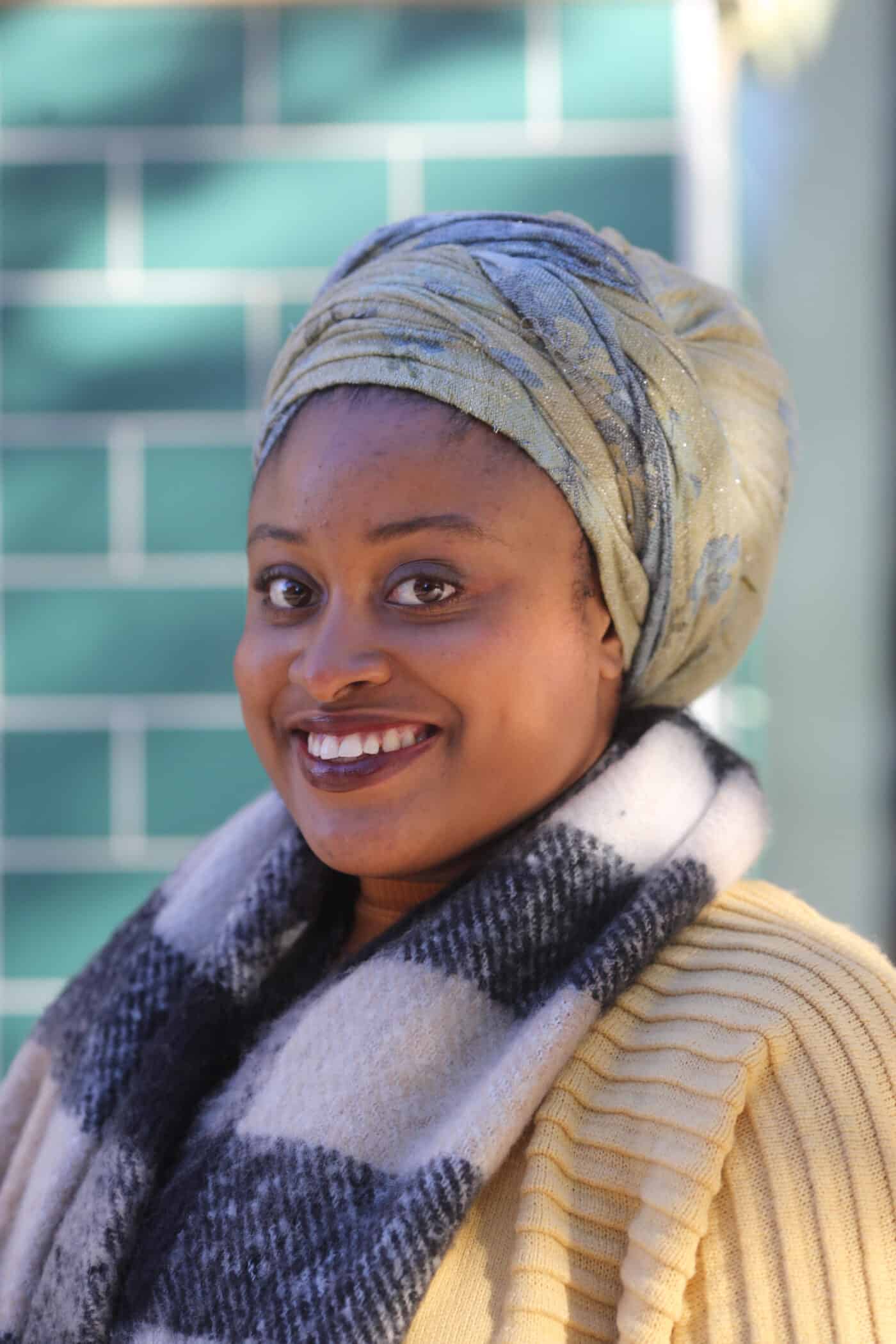 Britny, who is a black female with brown skin wearing a green and blue headwrap and a yellow cardigan.