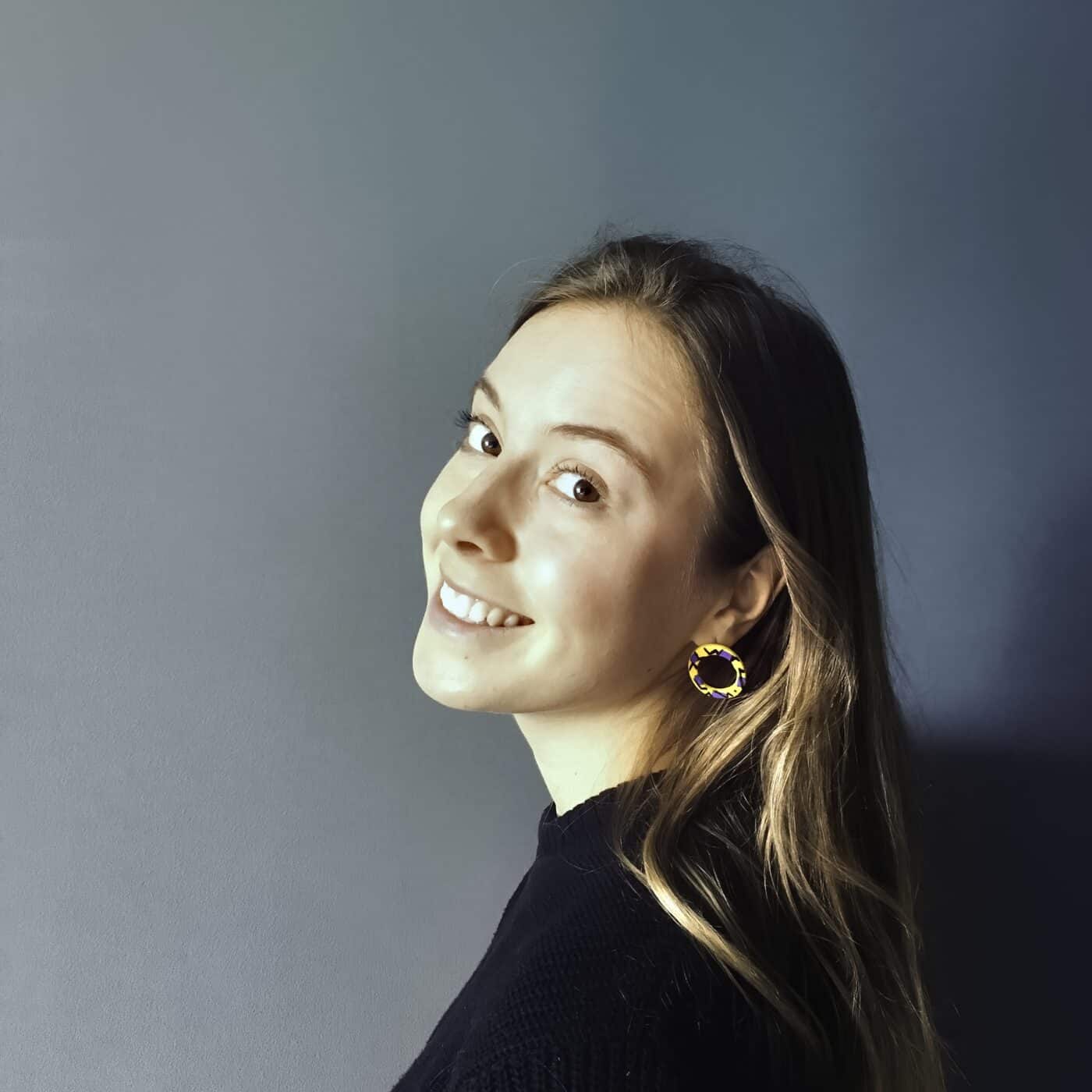Elspeth Wilson. A white person with long dark blonde hair smiles at the camera. They are facing side on and wearing a black jumper and yellow earrings and are against a dark grey wall.