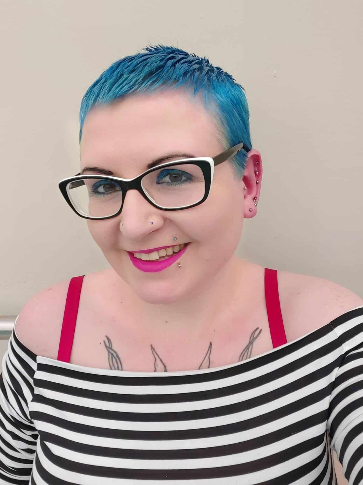 Gemma Lees. A white woman with short, blue hair, black plastic-rimmed glasses and pink lipstick, wearing a black and white stripy top.