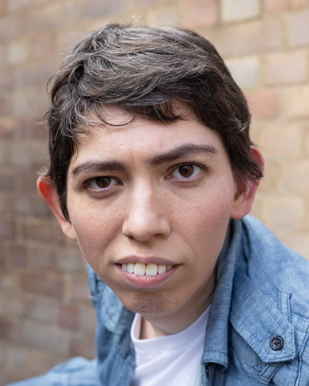 A headshot of Jessi, a white non-binary person with short brown hair and brown eyes, sitting in their powered wheelchair (out of shot) in front of a blurred brick wall. Jessi is wearing a light blue shirt over a white t-shirt, and gazing gently at the camera.