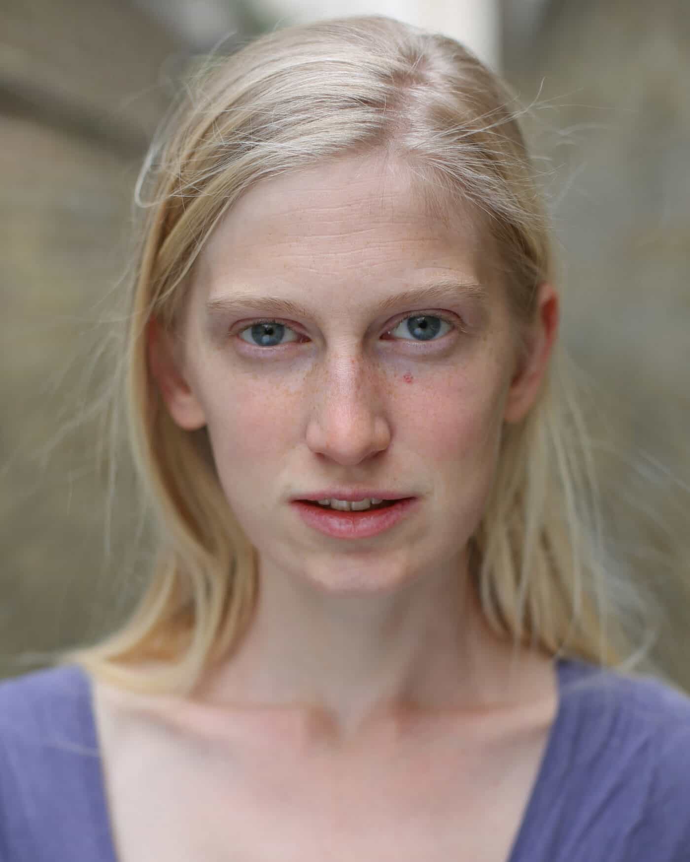 An image of Nell Hardy. A pale white woman in her early thirties with long blonde hair, blue eyes and freckles, wearing a pale purple top, smiles slightly in front of a blurred alleyway in the background.