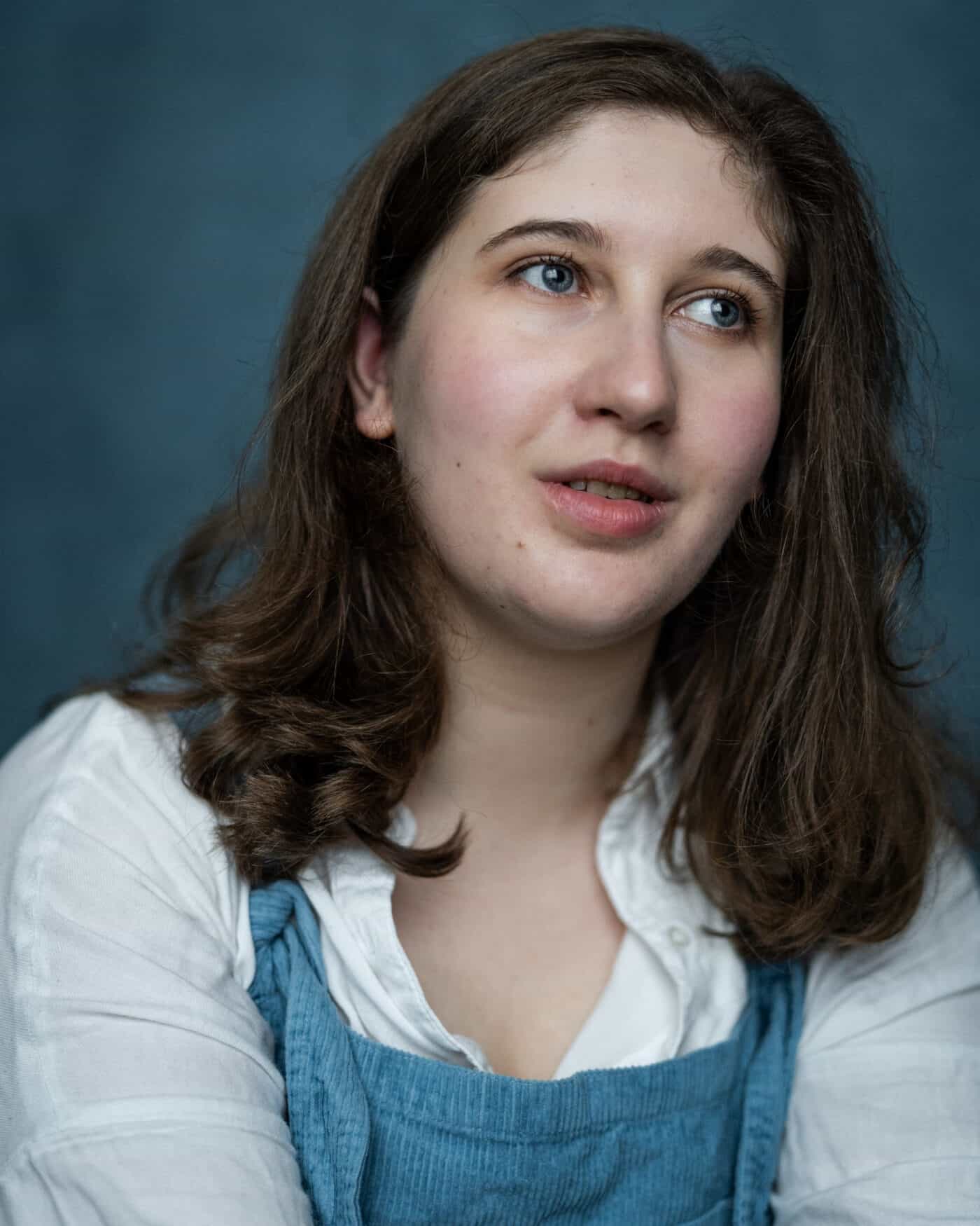Terri Donovan. Headshot of a young white person with shoulder length brown hair and grey eyes. They are wearing a white button down shirt and blue dungarees. They are looking away from the camera.
