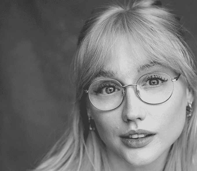 A portrait black and white photograph. Alice wears clear circlular glasses, she has a fringe and her hair is half up half down.
