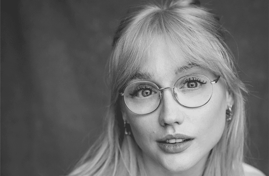 A portrait black and white photograph. Alice wears clear circlular glasses, she has a fringe and her hair is half up half down.