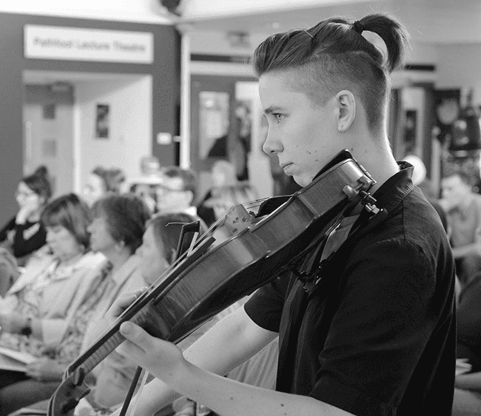 A black and white photo of Rufus. Rufus Elliot is pictured playing its viola, focusing on something out of shot. It is wearing a black short sleeved shirt, with its short hair tied up at the back.