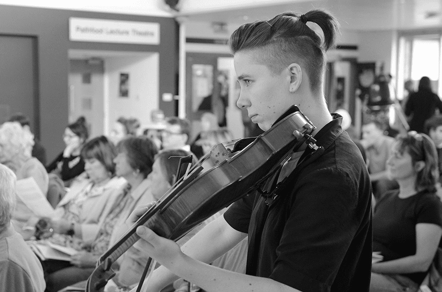 A black and white photo of Rufus. Rufus Elliot is pictured playing its viola, focusing on something out of shot. It is wearing a black short sleeved shirt, with its short hair tied up at the back.