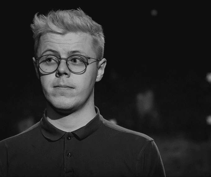 This is a black and white portrait. Tom is standing on stage, he is a caucasian boy with short-ish white hair. He is wearing round, plastic blue glasses and a black buttoned up polo shirt.