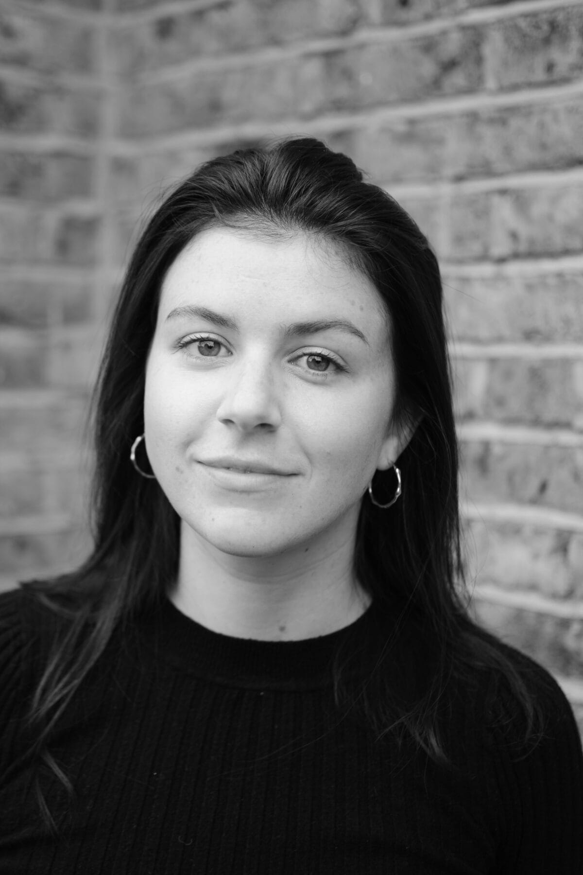 Black & White headshot of Caitlin Richards. Caitlin is a white woman with long brown hair and hazel eyes. She looks softly towards the camera. She is wearing a black jumper and small gold hoops. Behind her is a brick wall.