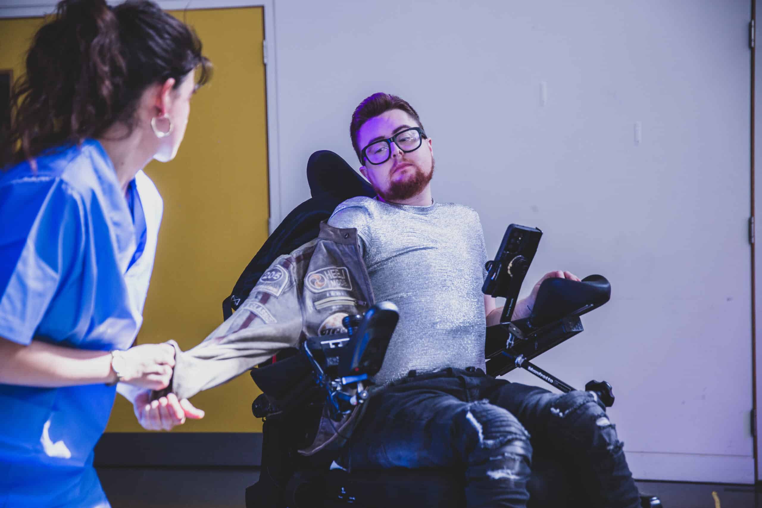 Jamie, a white person with dark red hair and beard, sits in an electric wheelchair, wearing ripped black jeans and a silver top. Sophie, a white woman with brown hair in blue scrubs is at the side of the image, pulling a leather jacket off Jamie's arm, as Jamie looks at her, camp and poised