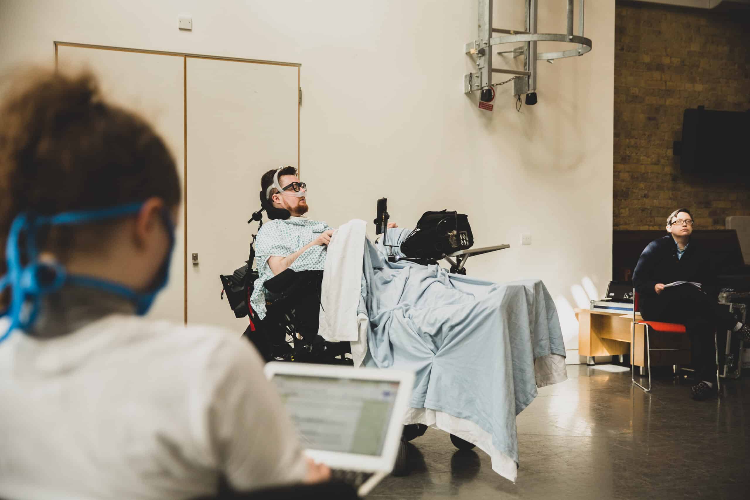 Jamie, a white person with dark red hair and beard, sits in an electric wheelchair, made to look like a hospital bed with blankets and sheets. They are wearing a ventilator mask. To the left, a person sits using a laptop, they have long brown hair, and are out of focus.
