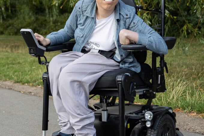 A full-length shot of Jessi, a white non-binary person with short brown hair and brown eyes, showing them sitting in their powered wheelchair, which is parked on a path in front of some green grass and trees. They are gazing gently at the camera. Their right hand is resting on their wheelchair control and their left is on their armrest. Jessi is wearing a light blue shirt over a white t-shirt with an obscured slogan, grey trousers and dark blue Converse shoes.