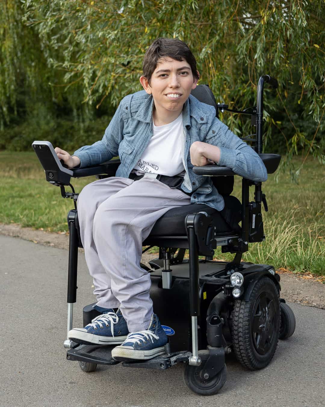 A full-length shot of Jessi, a white non-binary person with short brown hair and brown eyes, showing them sitting in their powered wheelchair, which is parked on a path in front of some green grass and trees. They are gazing gently at the camera. Their right hand is resting on their wheelchair control and their left is on their armrest. Jessi is wearing a light blue shirt over a white t-shirt with an obscured slogan, grey trousers and dark blue Converse shoes.