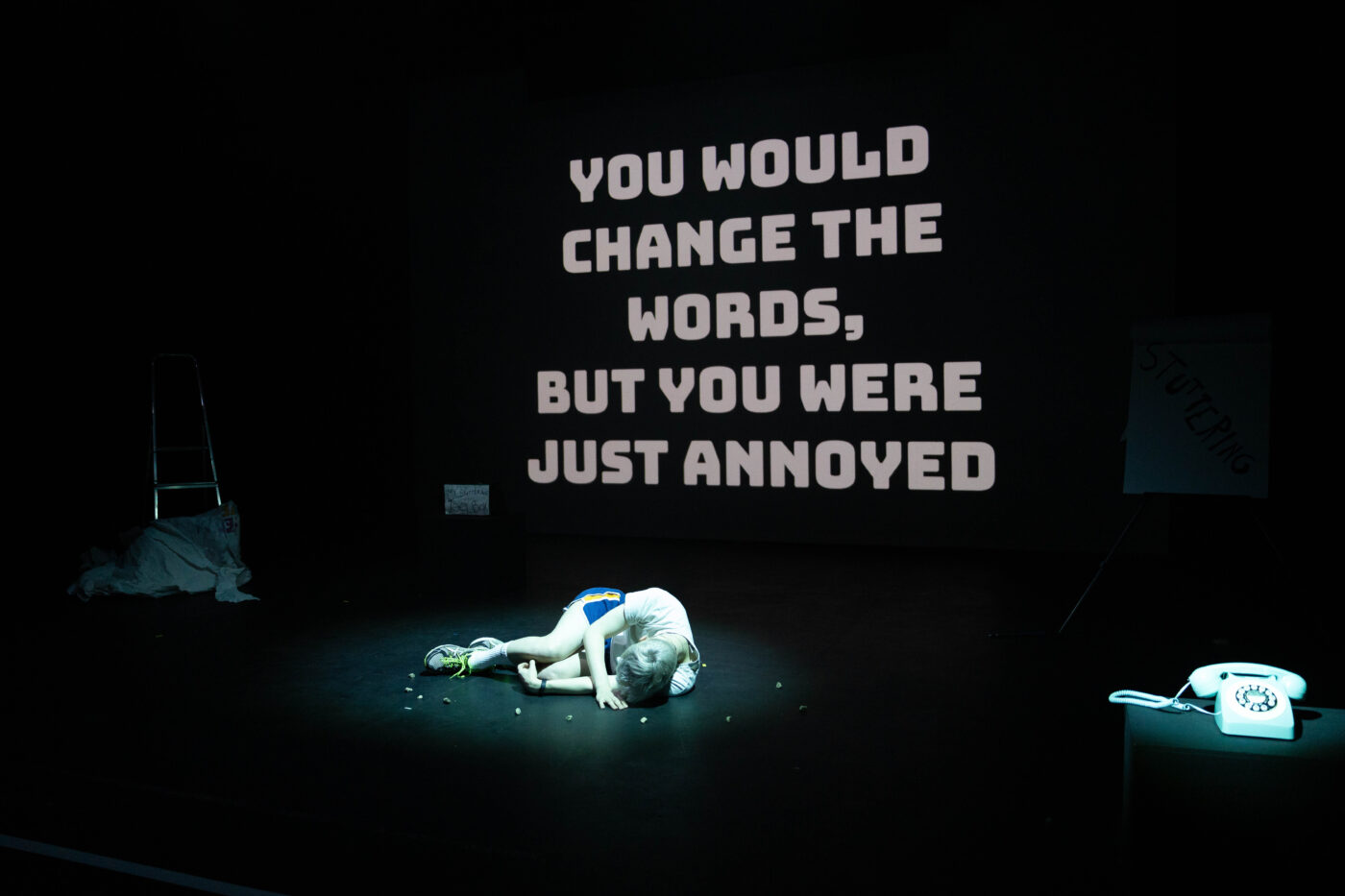 Oli - a white non-binary person - has bleached hair and is dressed in a white tshirt and blue gym shorts. They are curled up on the floor on a stage, surrounded by a circle of pebbles, and bathed in a cold spotlight. Behind them, the words 'you would change the words, but you were just annoyed' are projected onto a large screen. In the foreground, there is an old fashioned phone, also in a pool of cold light.
