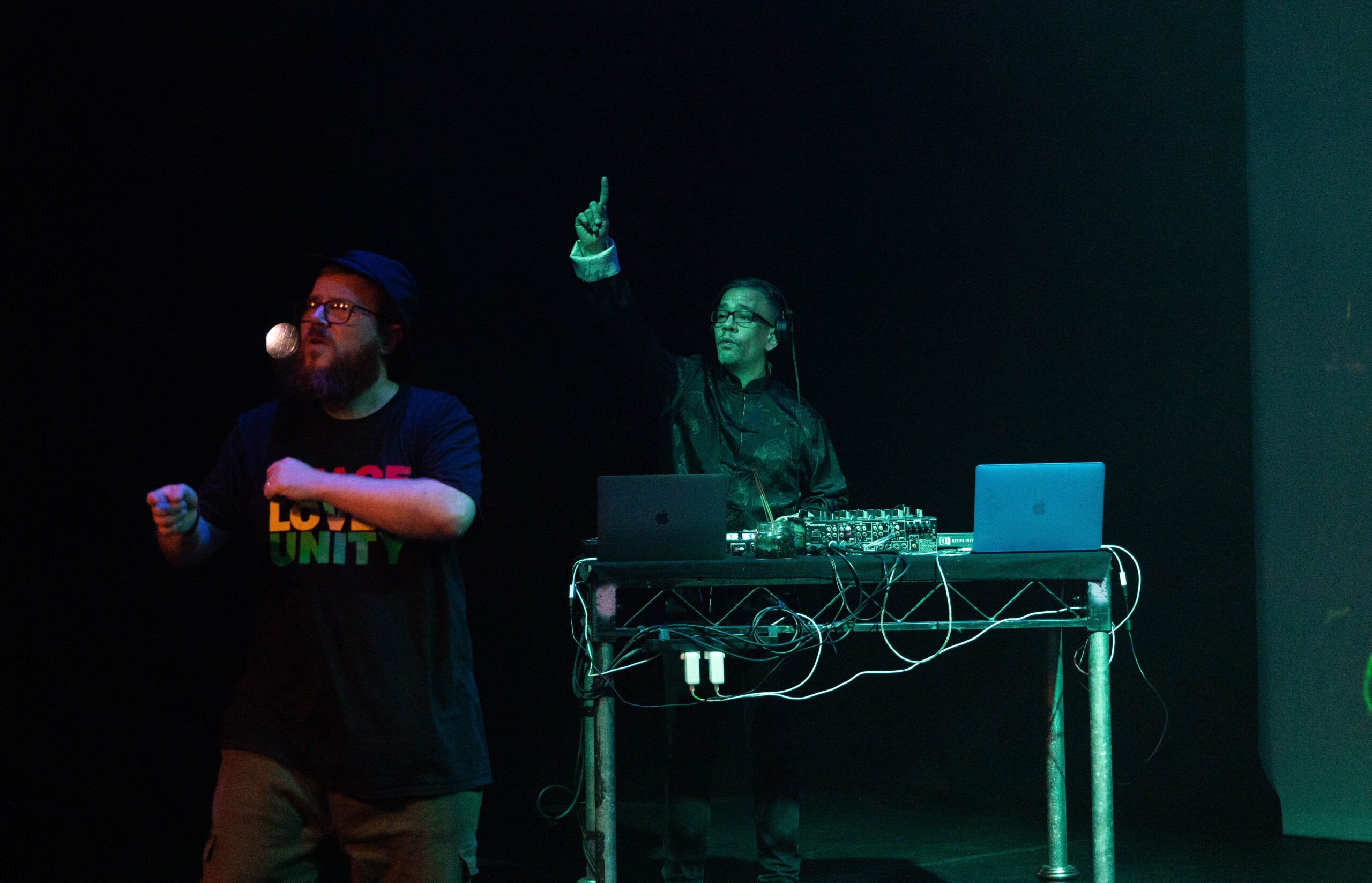 MC Geezer and Troi 'DJ Chinaman' Lee perform at the CRIPtic Pit Party in 2021. MC Geezer raps and signs on stage, while Troi Lee plays music from behind a desk, with his hand raised.