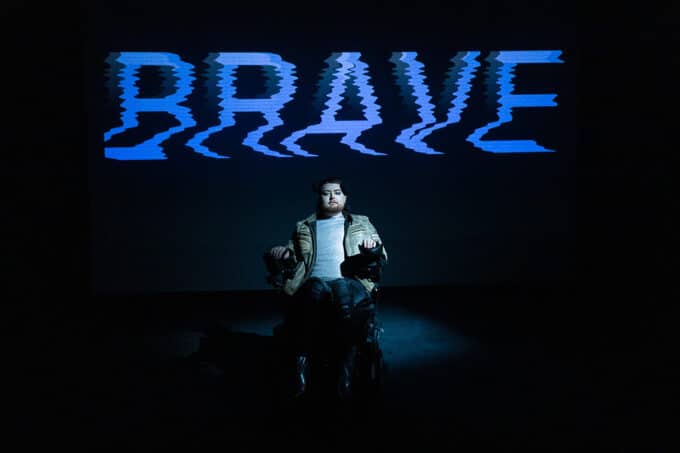 Jamie Hale performs NOT DYING. Jamie, white person with red hair, wears a silver shirt, a beige leather jacket and grey jeans. They stare into the camera as the word 'Brave' is projected and glitches behind them.