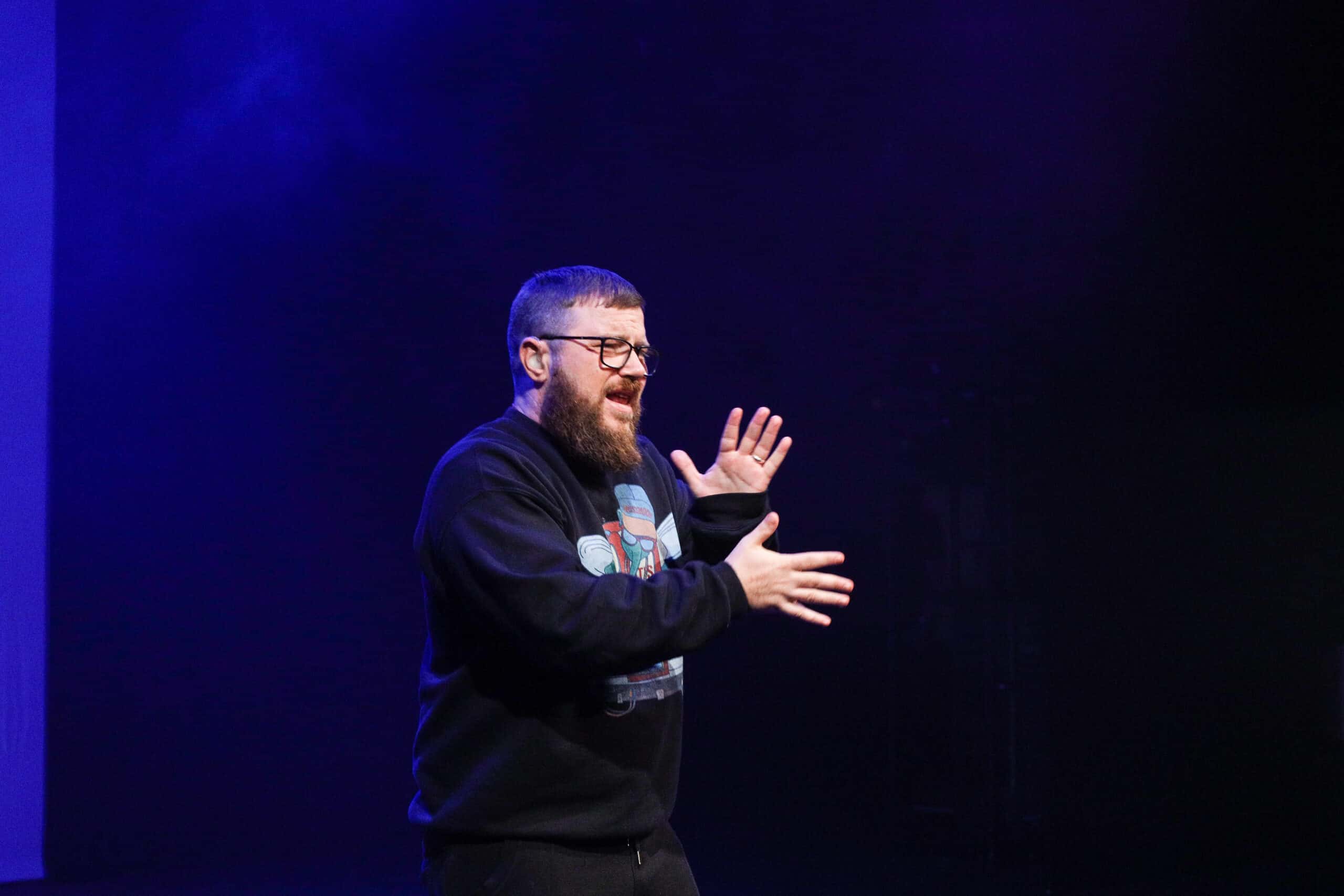 MC Geezer a white man with short brown hair and beard. He has black glasses, and is wearing a blue jumper with a large logo in the centre. He signs on stage at the Barbican.