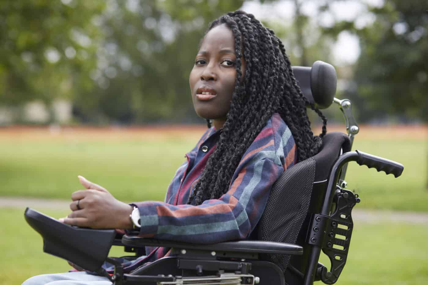 Jacqui is a black woman with black and slightly grey waist-length twists, She is wearing a vertically striped shirt. She is sitting in her powered wheelchair sideways facing, Jacqui staring straight into the camera with a curious look on her face. The background is greenery not in focus.