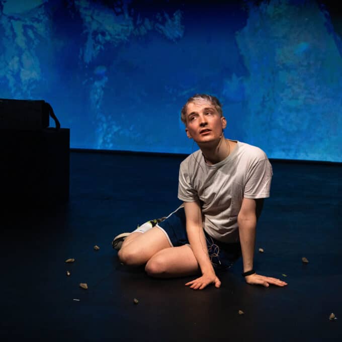 Oli - a white non-binary person - has bleached hair and is dressed in a white tshirt and blue gym shorts. Oli sits on stage, with a blue-washed projection behind them. They have a focused expression and their mouth is slightly open.