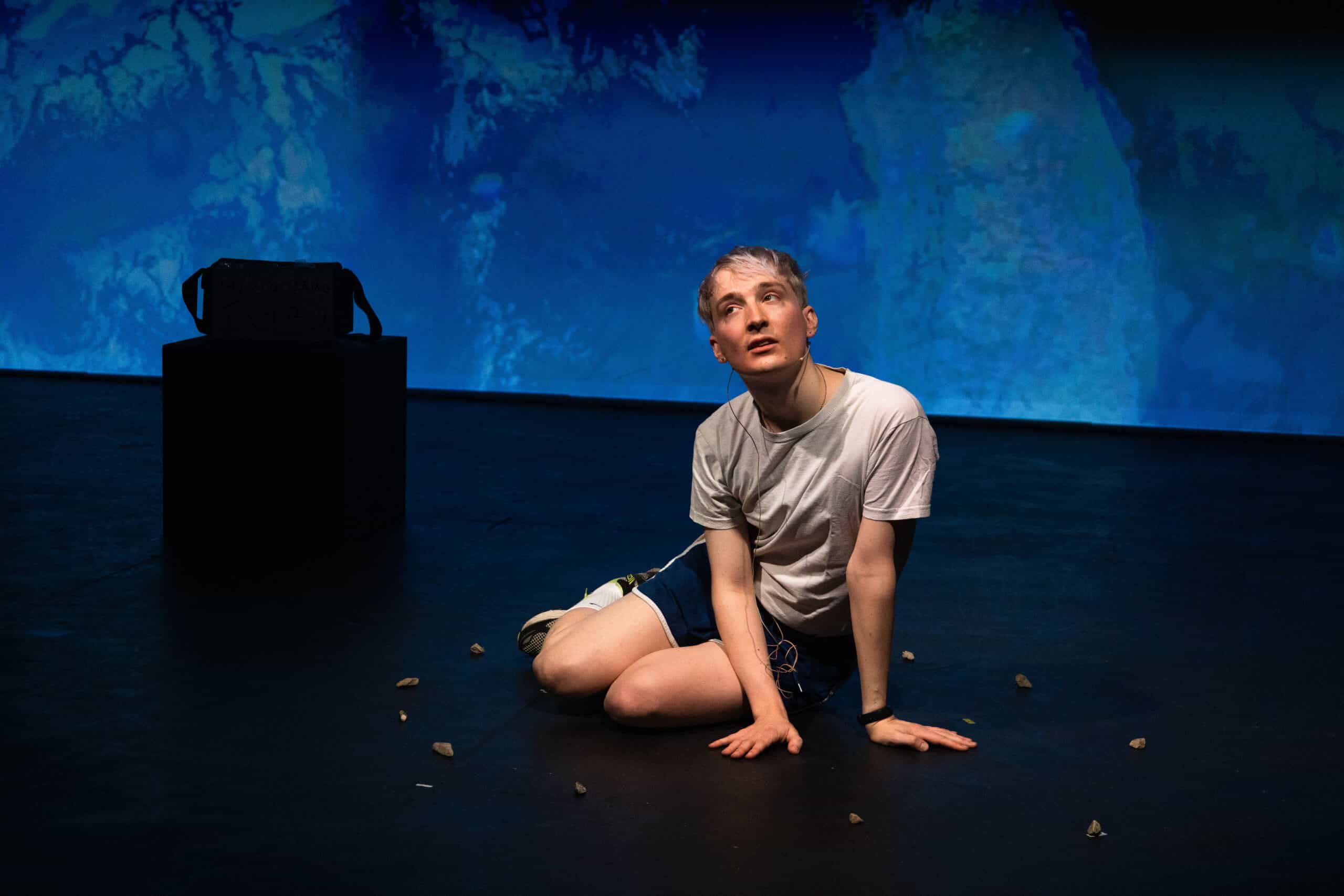 Oli - a white non-binary person - has bleached hair and is dressed in a white tshirt and blue gym shorts. Oli sits on stage, with a blue-washed projection behind them. They have a focused expression and their mouth is slightly open.