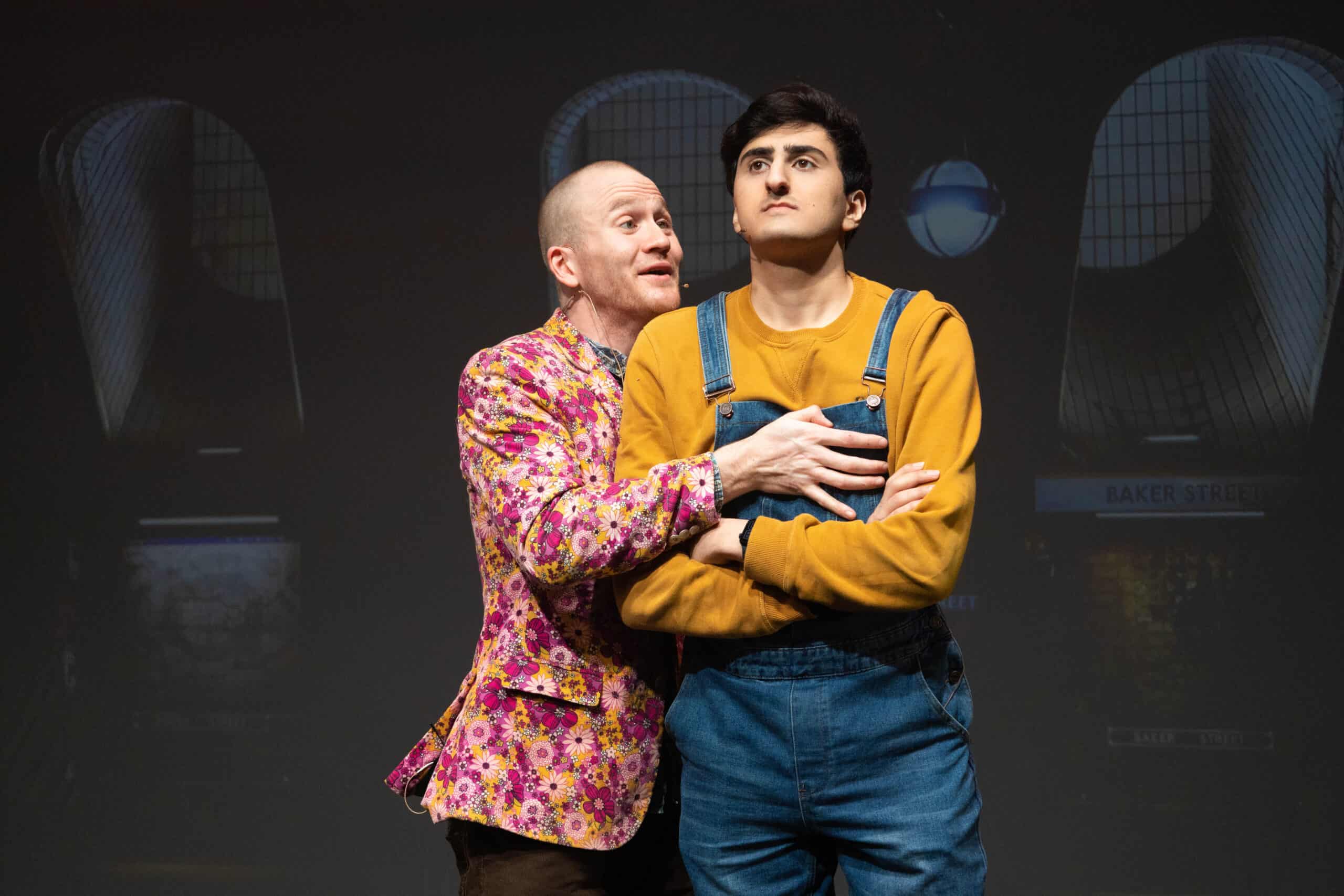 Sam Brewer & Aarian Mehrabani (Sam, a white man with closely shaved blonde hair wears a pink and yellow floral blazer and grasps a very resolute Aarian. Aarian is a Middle Eastern man with slim build, clean shaven with short jet black quiffed hair. He is wearing a yellow jumper and denim dungarees).