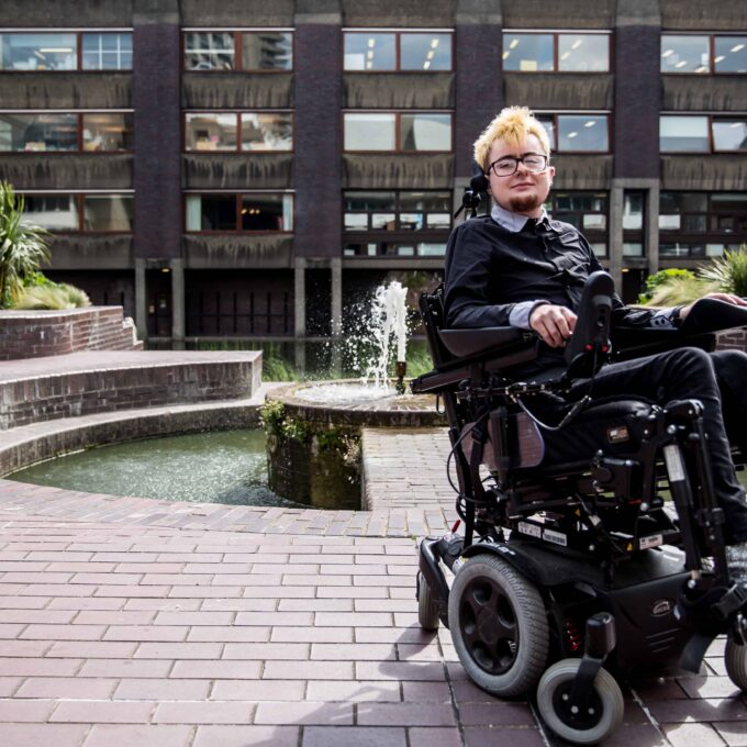 Jamie Hale, a white person in a black electric wheelchair, wearing all black, with silver boots, and beach-blonde hair, in front of the brutalist Barbican Centre