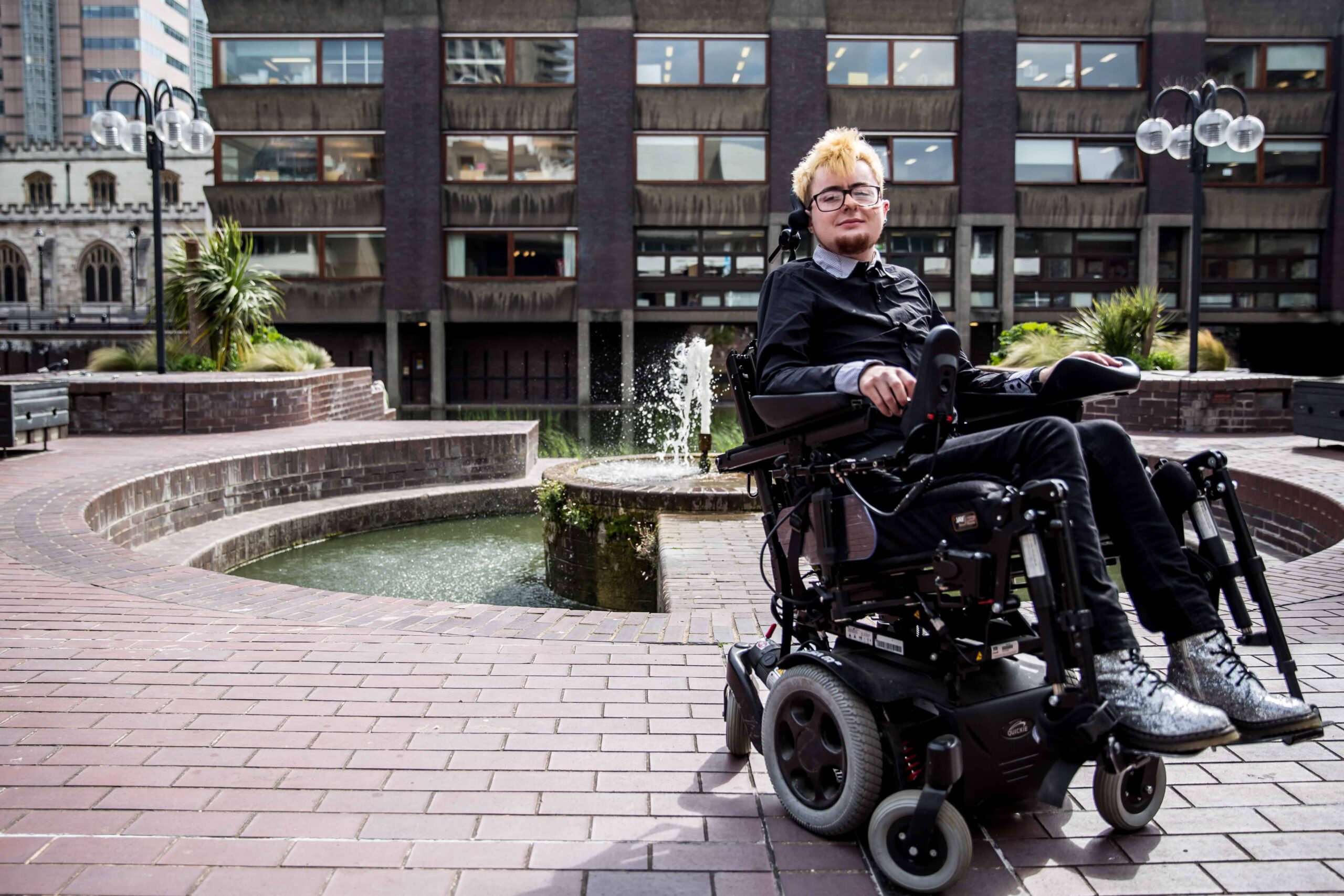 Jamie Hale, a white person in a black electric wheelchair, wearing all black, with silver boots, and beach-blonde hair, in front of the brutalist Barbican Centre