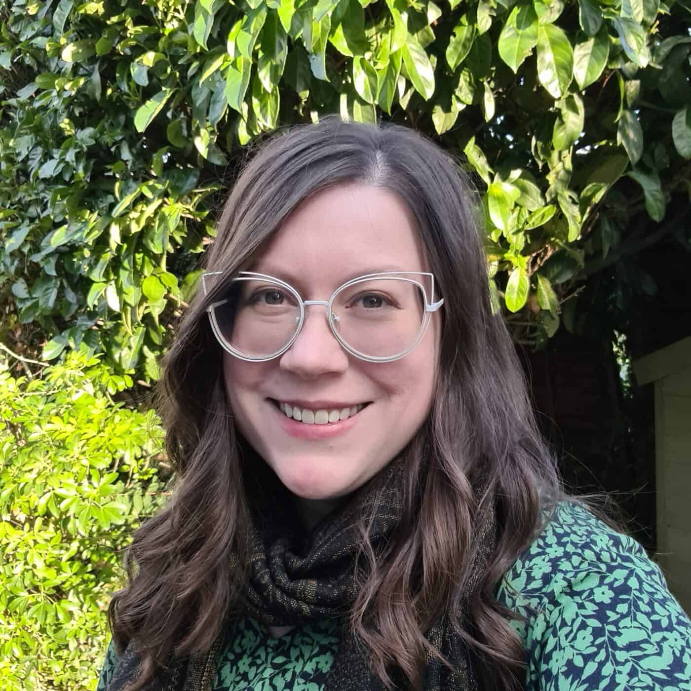 Alli Smith Headshot. A white woman with brown hair and white glasses is smiles in front of a background of leaves.