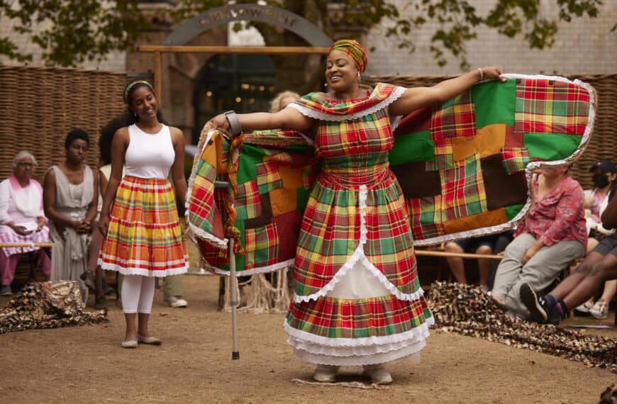 Vibrant outdoor scene, the focal point of which is a black female with brown skin wearing a dress featuring a green, red and yellow checked pattern, with a patchwork cape spread out to resemble wings.