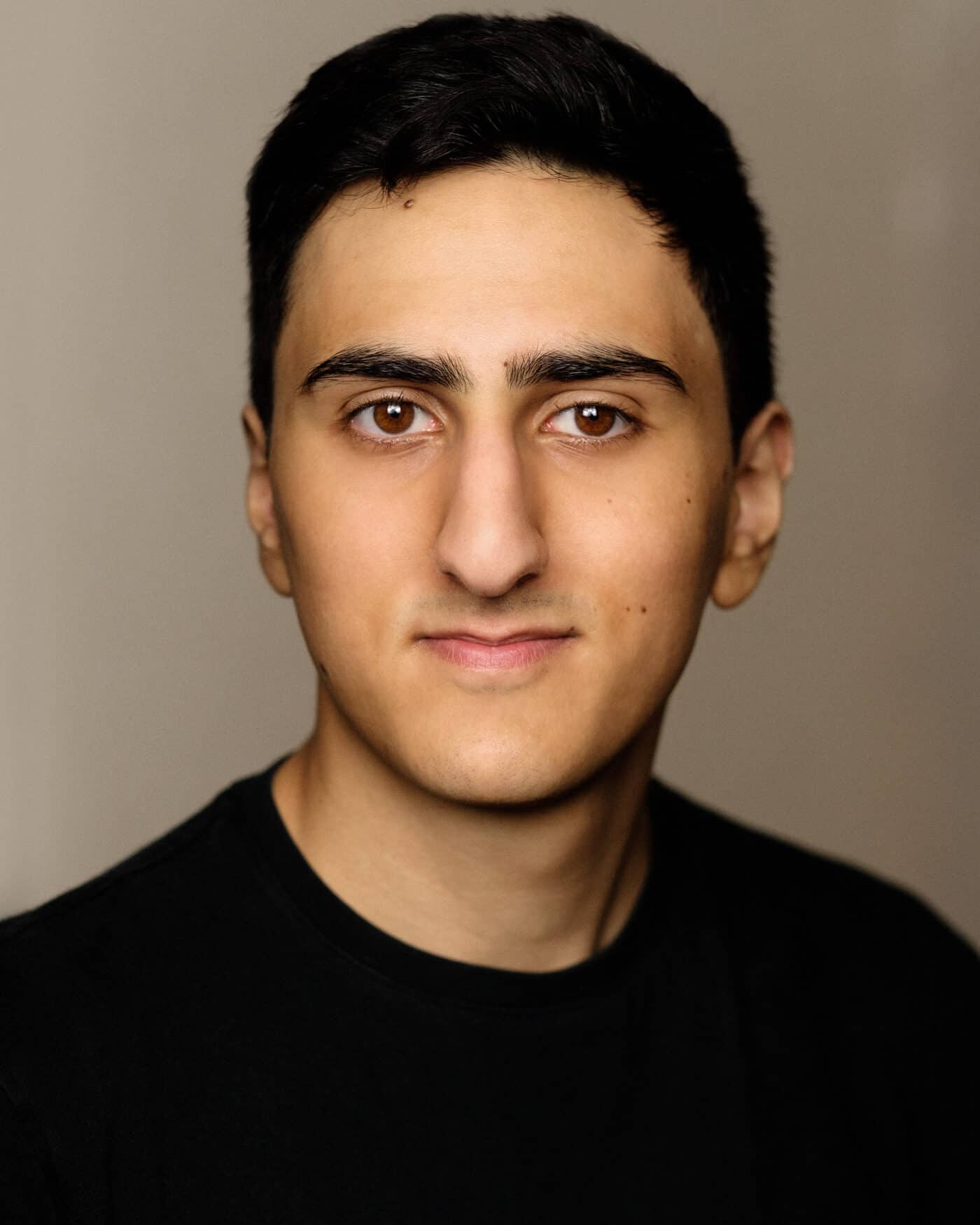 A head shot of Aarian Mehrabani. A clean shaven Middle Eastern Man in his early 20s looking directly at the camera. He has thick black hair and is wearing a black T-Shirt. 
