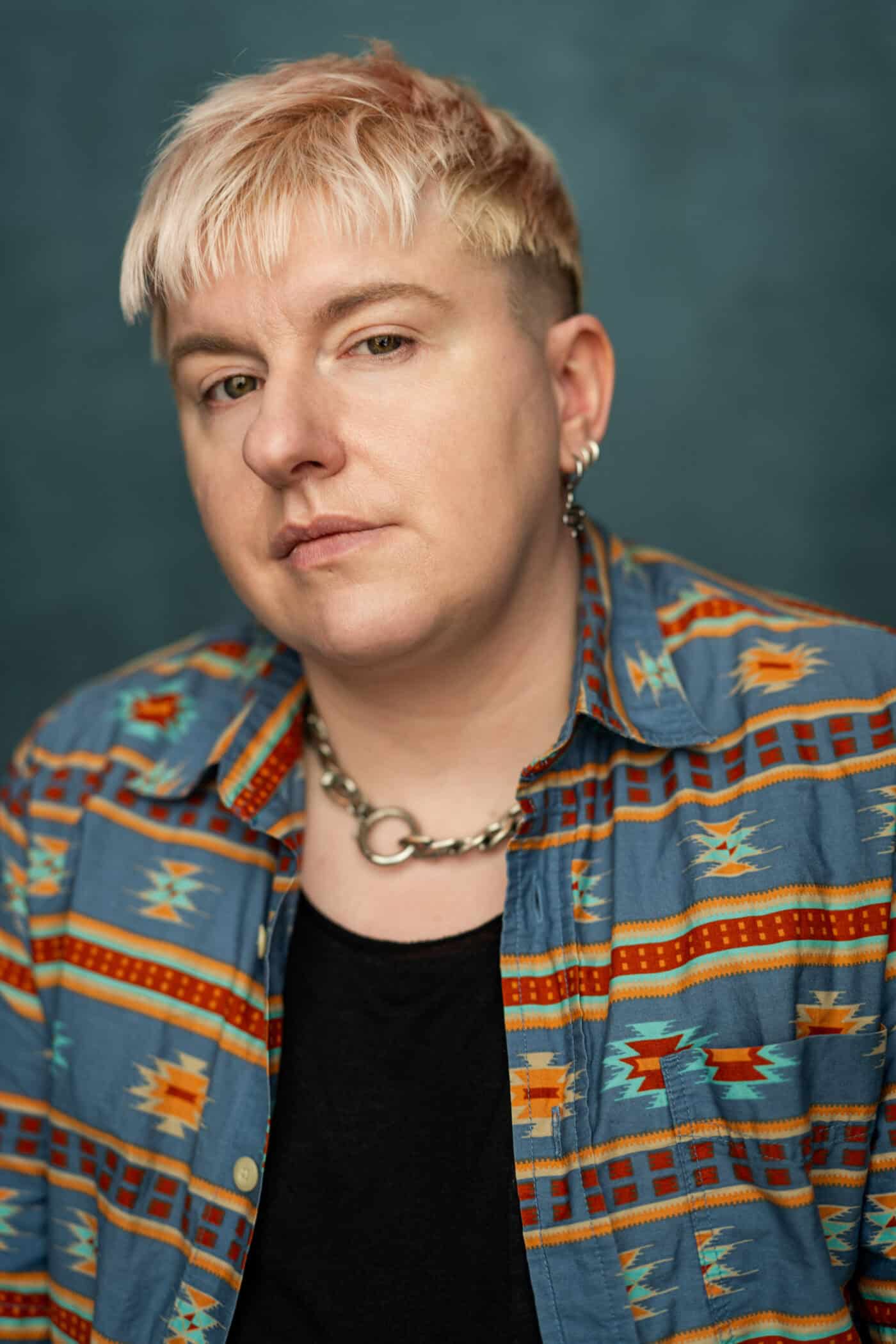 A headshot of a white person with short, very pale pink hair, wearing a geometric patterned shirt, a black undershirt and silver jewellery.