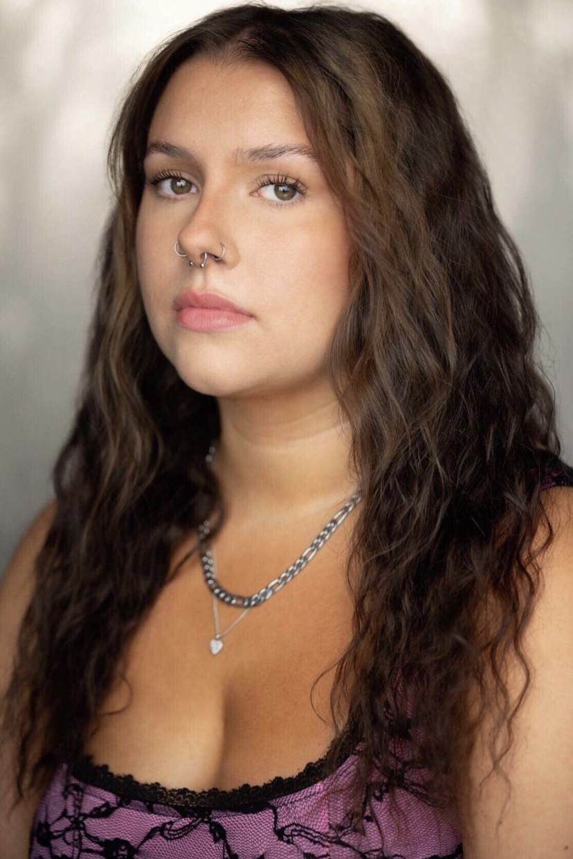 Katherine Castellucci's headshot. A white woman with long, dark brown, wavy hair is wearing three silver hoop nose rings, and two silver necklaces. One necklace is a thick chain, and the other is a thin chain with a silver heart pendant. She is wearing a hot pink, strappy top with a black lace overlay. She is looking at the camera with a stern expression.