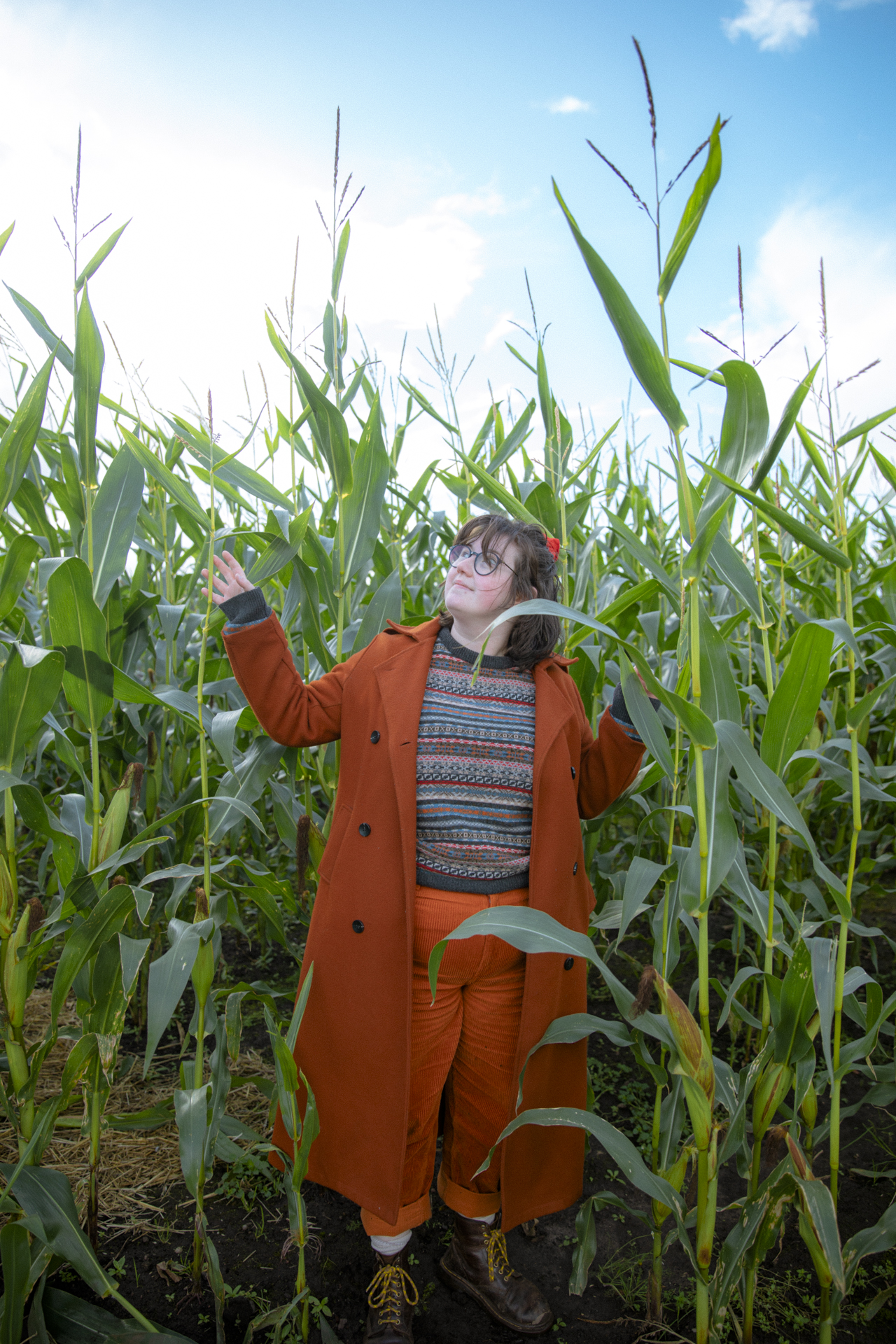 Meg, a white person in their twenties with brown hair and glasses, is stood in a corn field below a blue sky. They’re facing the camera but looking up to their right with arms in the air. They’re wearing a long brown coat, orange trousers and a patterned jumper.