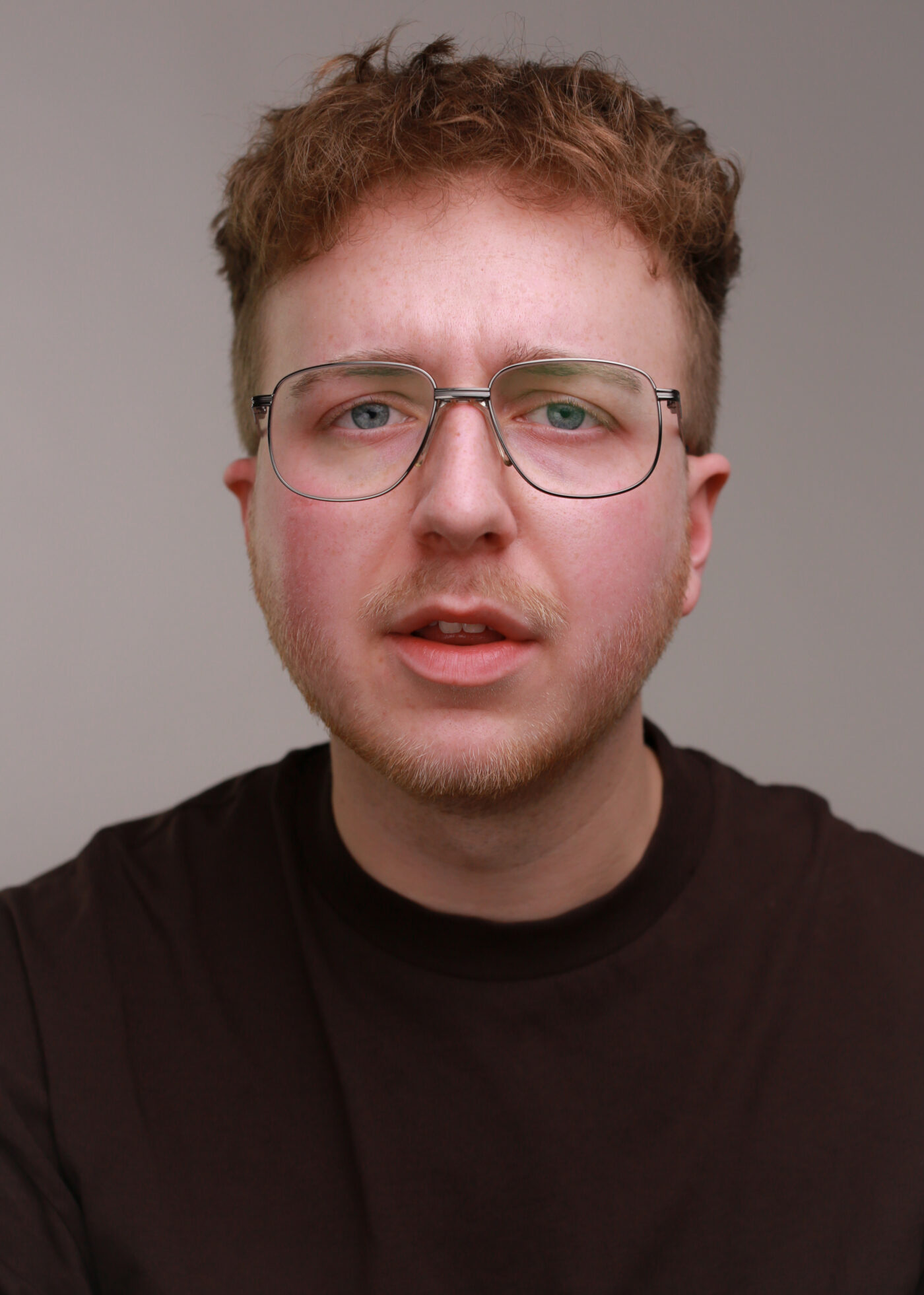 The image is a portrait of a person wearing metal
framed glasses. The person is indoors. They have short blonde curly hair, a high neck brown t-shirt,
freckles and a short moustache. Their mouth is slightly open.